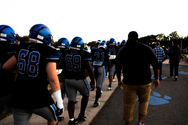 Gruppe von Füßballspielern in Helmen und Stiefeln, die über eine Straße mit Bäumen und einem klaren blauen Himmel im Hintergrund gehen.
