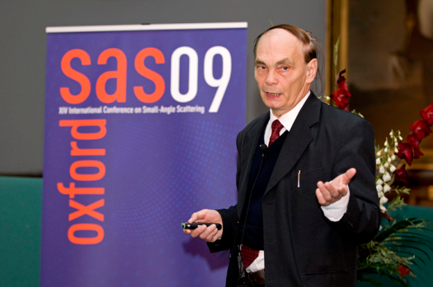 Ein älterer Mann in einem Anzug hält ein Objekt und spricht, mit einem Banner, einer Wand, einem Foto Rahmen und Blumen im Hintergrund.