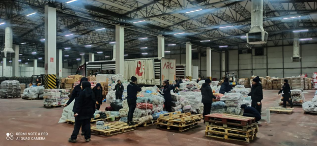 Eine Gruppe von Menschen steht um ein großes Lagerhaus herum, das mit Food-Tüten, Holzpaletten, Containern, Pfählen, einem Schild, einer Wand, einem Dach mit Deckenlampen und Metallpfählen sowie einem Wasserzeichen auf der linken Seite gefüllt ist und scheinbar ein Food-Verteilzentrum in Dubai ist.