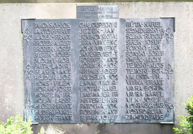 Ein Steinplakette an einer Wand mit eingraviertem Text, umgeben von Pflanzen unten, wahrscheinlich ein Denkmal für Holocaust-Opfer.