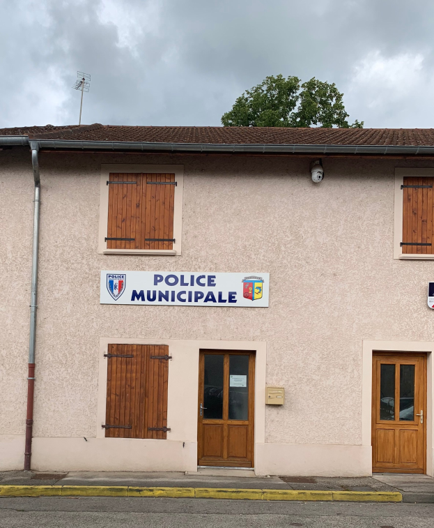 Gebäude der Police municipale mit einem Schild, Fenstern, Türen, einer Namensplakette, einem Rohr, einem Baum und einem bewölkten Himmel im Hintergrund.