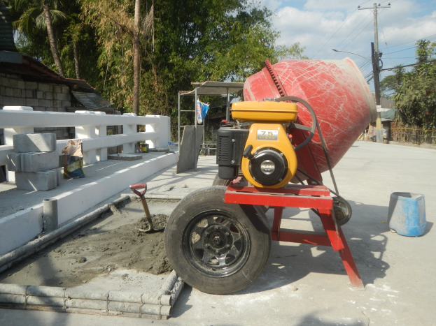 Betong-Mischer auf einem Zement-Lkw umgeben von Baustoffen und einem Haus mit Bäumen unter einem bewölkten Himmel.