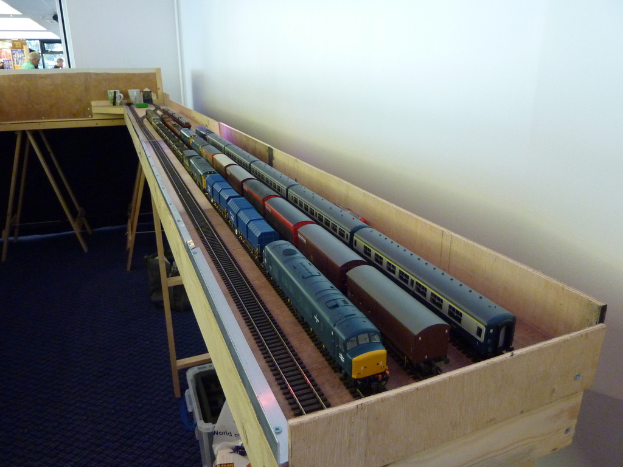 Ein auf einem Tisch mit Schienen, Wand und Boden im Hintergrund stehendes Spielzeughaus, mit Menschen in Kleidung im Hintergrund.