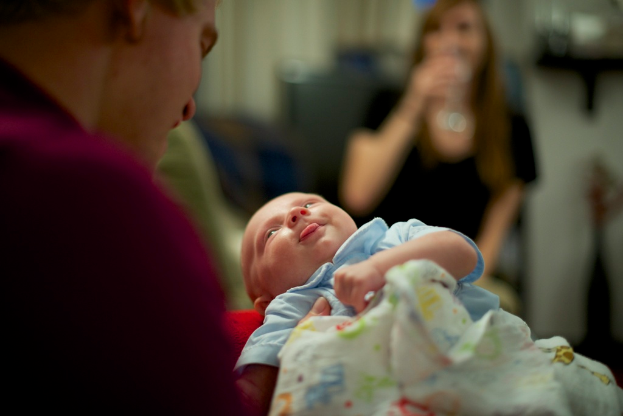 Ein Mann hält ein Baby in den Händen, vor ihm eine Frau, mit einem unscharfen Hintergrund.