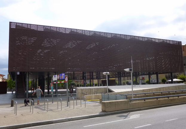 Großes modernes Gebäude mit zahlreichen Fenstern, das als neuer Eingang zum Stadtzentrum dient, umgeben von Straßeninfrastruktur, Fahrzeugen, Fußgängern und Bäumen unter einem bewölkten Himmel.