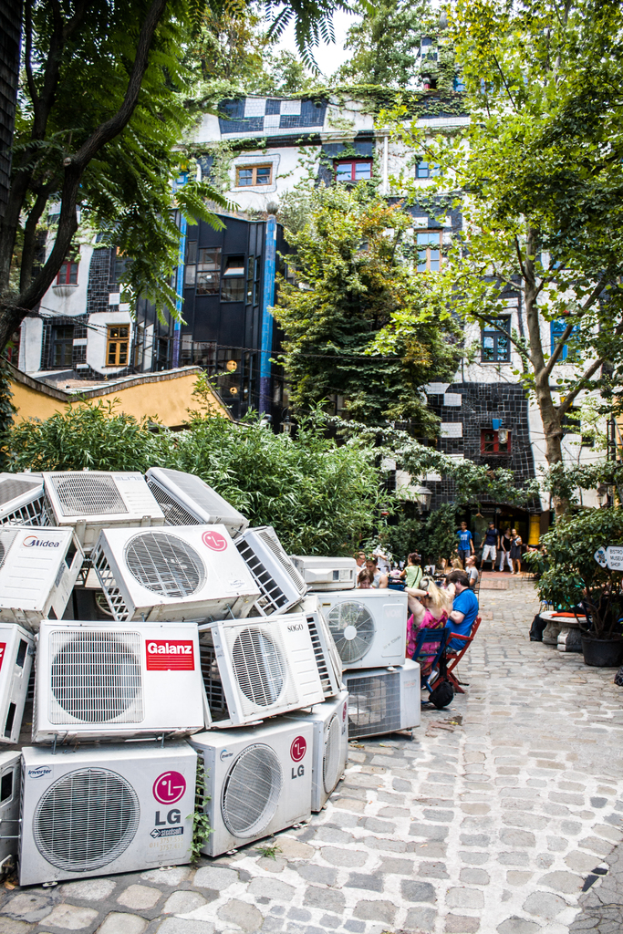 Luftkühler am Boden links unten, eine Straße rechts daneben, Menschen auf Stühlen neben der Straße sitzen, mit Gebäuden, Bäumen und Pflanzen oben.