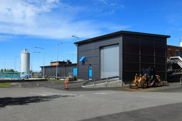 Großes Gebäude mit einem Bagger davor, umgeben von Straßeninfrastruktur und Fahrzeugen, unter einem bewölkten Himmel, als neue Industriepark-Anlage gekennzeichnet.