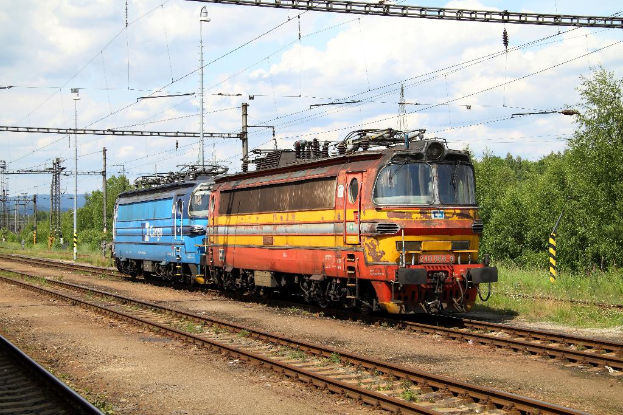 Ein Zugmaschine auf Schienen mit Strommasten, Kabeln, Bäumen, Pflanzen und Wolken am Himmel.