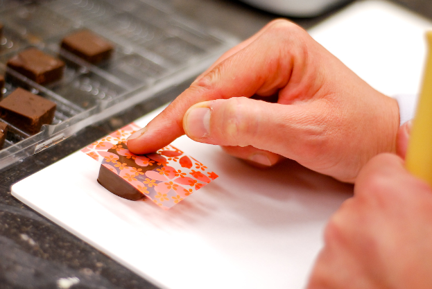 Eine Hand platziert eine dekorative Abdeckung auf einer Schokolade, mit weiteren Schokoladen auf einem Tablett links und einem Tisch darunter.