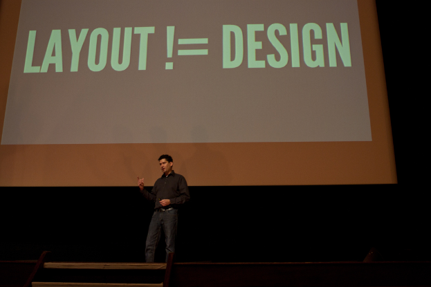 Eine Person steht auf einer Bühne und spricht, mit einem Bildschirm im Hintergrund, der Text anzeigt.