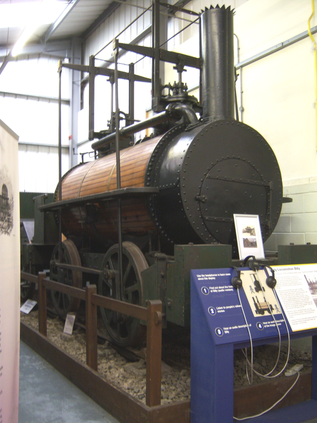 Alte Dampflokomotive in einem Museum ausgestellt, umgeben von Informationsschildern, einem Zaun und Deckenbeleuchtung.