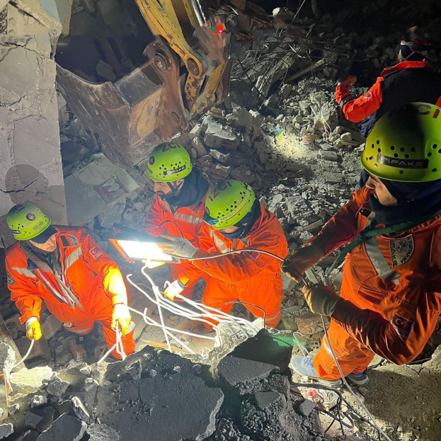 Gruppe von Rettungskräften in orangefarbenen Uniformen und grünen Helmen beim Beseitigen von Schutt von einem eingestürzten Gebäude mit einem Kran im Hintergrund.