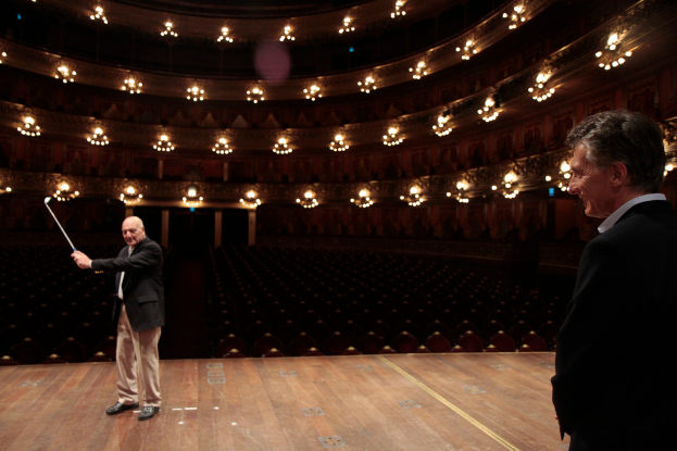 Ein älterer Mann in einem Anzug hält einen Golfschläger auf der Bühne, während ein anderer älterer Mann rechts von ihm steht, unter Deckenlampen mit vielen Sitzplätzen darunter.