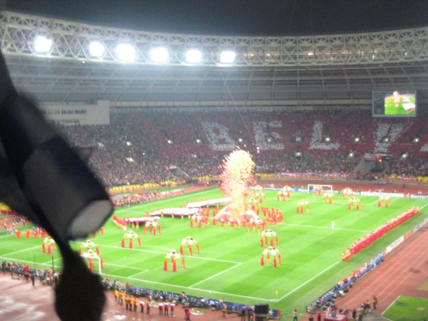 Ein Stadion mit Flutlicht, Eisengittern, Zuschauern, stehenden Individuen, Werbetafeln, Anzeigetafeln, einer Sicherheitskamera und verschiedenen Dekorationen einschließlich Flaggen.