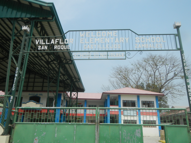 Der Eingang der Villaflor-Grundschule in San Roque mit einem Tor mit Text darauf, umgeben von Gebäuden mit Fenstern, Geländern und einem Schuppen, mit Bäumen und einem klaren blauen Himmel im Hintergrund.