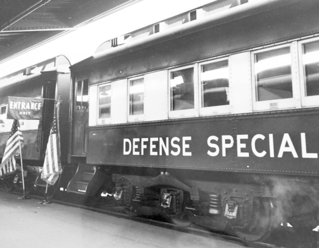 Schwarz-weißes Foto eines Zuges mit der Aufschrift "Defense Special" auf den Eisenbahnschienen mit zwei Fahnen links und einer Decke darüber.