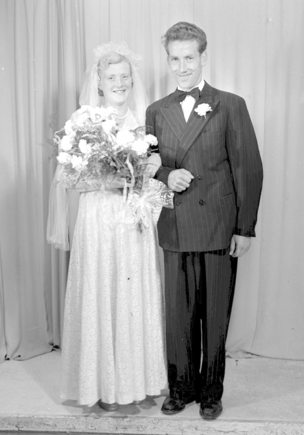 Ein Mann und eine Frau stehen nebeneinander auf einer Bühne, lächeln und halten ein Blumenbouquet, mit einem Vorhang dahinter.