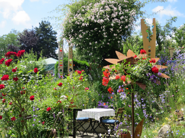 Ein Garten mit bunten Blumen und Pflanzen, mit einem Tisch mit einer Decke und Stühlen, vor Bäumen und einem bewölkten Himmel, auf dem Chelsea Flower Show.