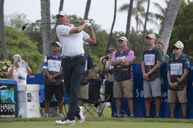 Ein Mann in einem weißen T-Shirt, schwarzen Hosen und einer blauen Mütze schwingt einen Golfschläger auf einem saftig grünen Feld, mit Zuschauern und Fotografen hinter ihm unter einem klaren blauen Himmel mit Bäumen im Hintergrund.