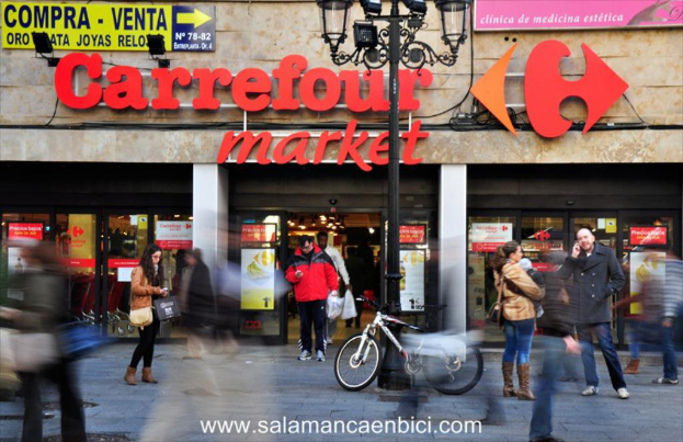 Ein belebter Einkaufsmarkt mit Menschen vor einem Einkaufszentrum, einem schwarzen Pfahl mit Lichtern, einem Fahrrad daneben und einem Wegweiser an der Wand.