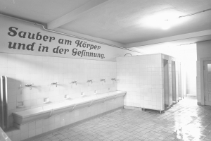 Schwarz-weißes Foto einer öffentlichen Toilette mit Fliesenboden, Text an der Wand, mehreren Wasserhähnen, einer Tür und einer Deckenlampe.