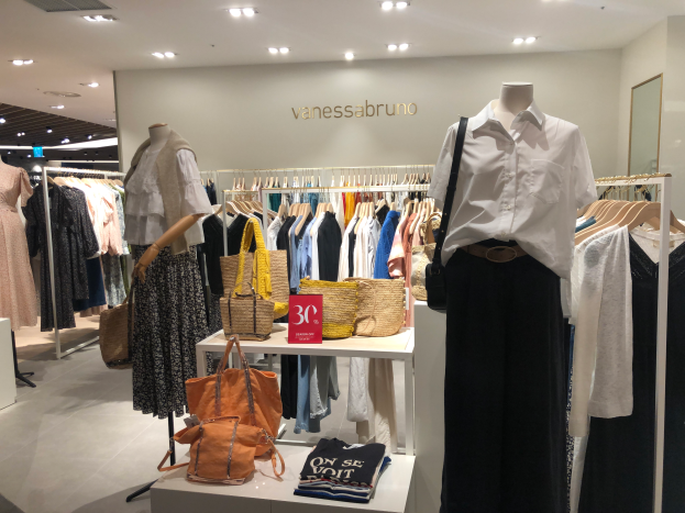 Ein Vanessa Bruno Geschäft in Singapur mit Schaufensterpuppen in Kleidern, hängenden Kleidern, Taschen, einer Tafel an der Wand, einem Spiegel auf der rechten Seite und Deckenlampen.