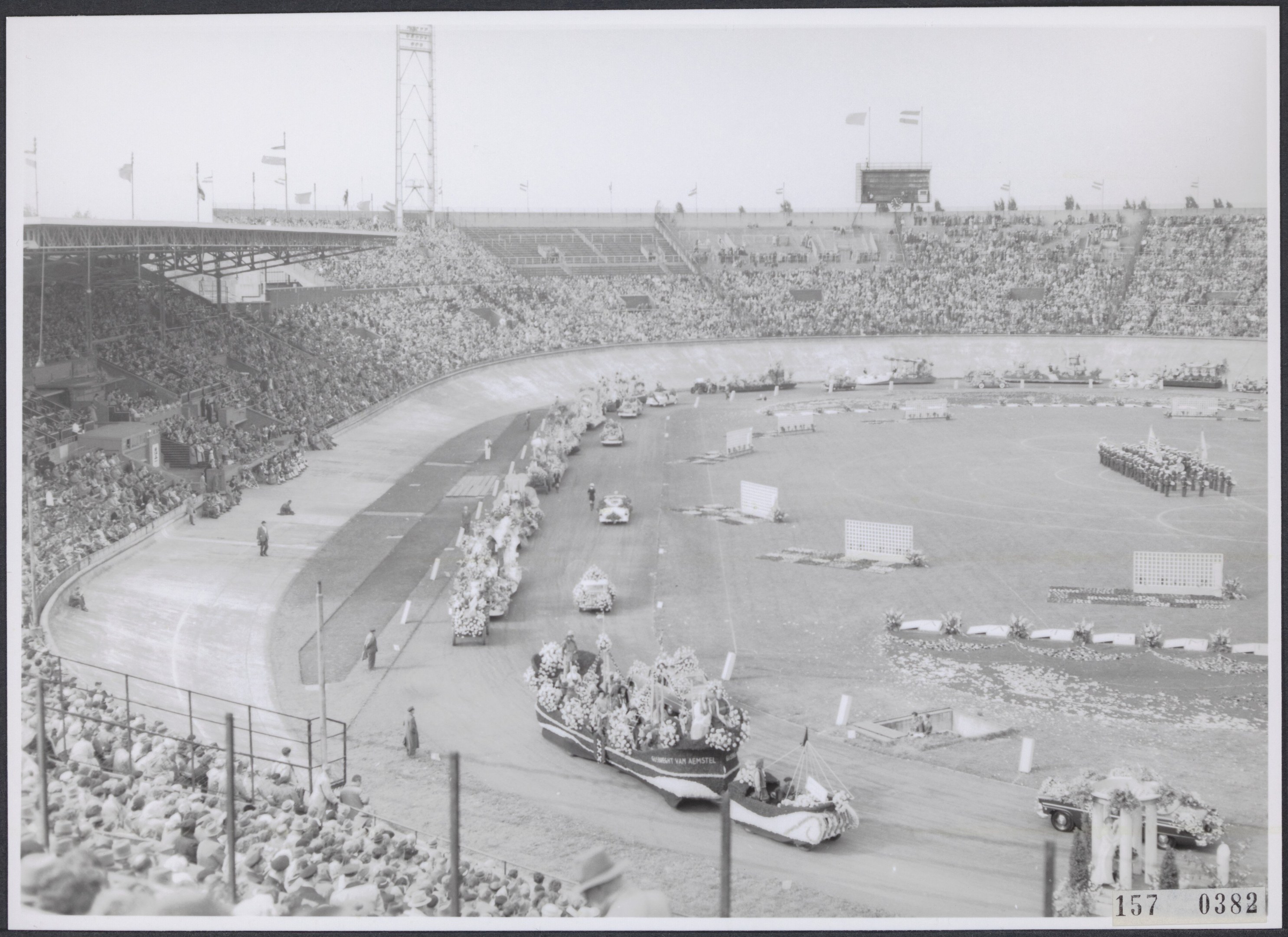 Schwarzes und weißes Foto einer Rennstrecke mit Zuschauern in einem Stadion, Fahrzeugen auf der Straße und Fahnen im Hintergrund.