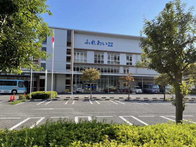 Ein großes Gebäude mit Fenstern und Text, das als Hauptquartier des japanischen Busdienstzentrums dient, mit zahlreichen parkenden Autos davor, umgeben von Pflanzen, Bäumen, Pfählen, Flaggen und Verkehrskegeln, unter einem klaren blauen Himmel.