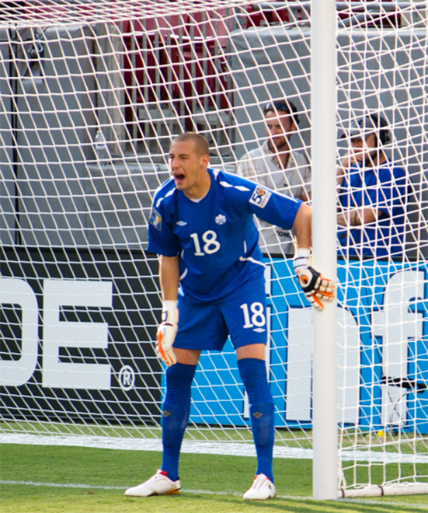 Ein Fußballspieler steht vor einem Tor mit ausgestreckten Armen, trägt eine Sportuniform und Schuhe, mit drei Personen, einem Netz und einer Tafel mit Schrift im Hintergrund.