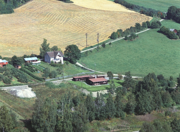 Luftaufnahme eines Bauernhofs in einem grünen Feld, umgeben von Bäumen, Häusern, Masten und einer gewundenen Straße.