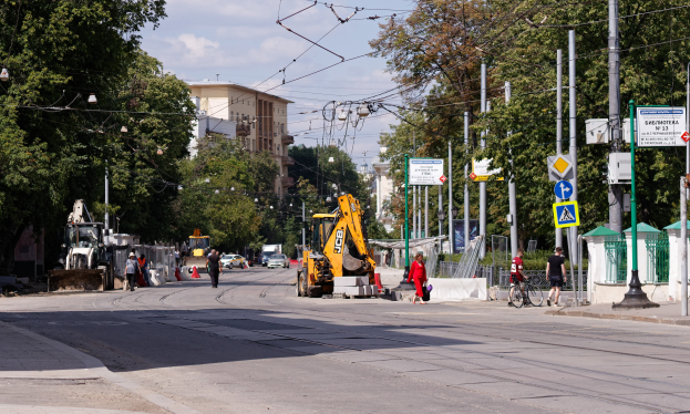 Eine Stadtstraße mit einer Baustelle in der Mitte, mit Fahrzeugen, Fußgängern, einem Radfahrer, Verkehrsleitkegeln, Pfählen, Hinweistafeln, Strommasten mit Drähten, Bäumen, Gebäuden mit Fenstern und einem bewölkten Himmel im Hintergrund.