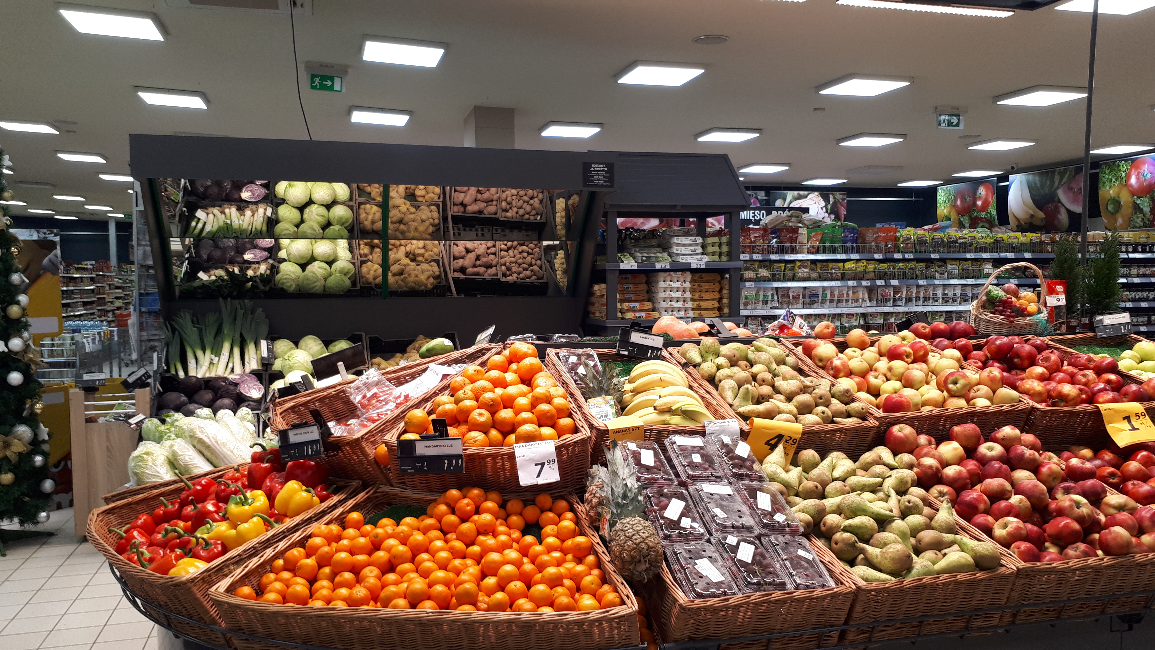 Ein Geschäft mit Körben voller Obst und Gemüse, Preisschildern, einem geschmückten Weihnachtsbaum links und Regalen im Hintergrund.