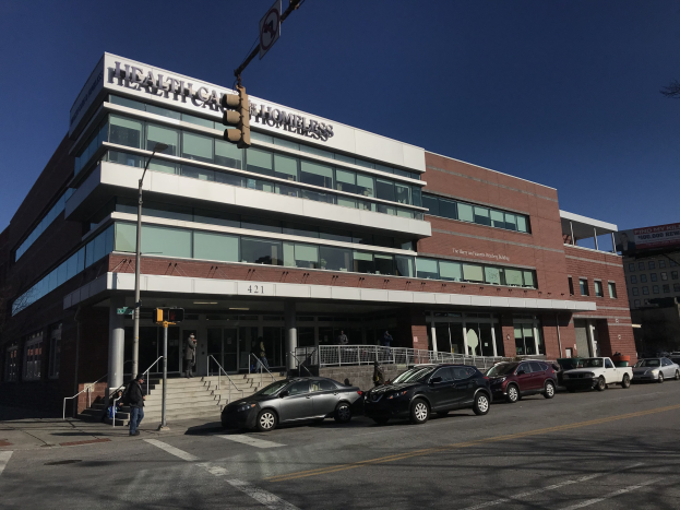 Ein großes Gebäude, das als New York City Health Care Homeless Center identifiziert wurde, mit parkenden Autos davor, Straßenlaternen, Verkehrsschildern, Menschen, die auf dem Gehweg gehen, Geländern, Treppen, Bäumen und einem klaren blauen Himmel.