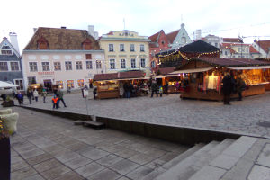 Ein lebendiger Weihnachtsmarkt in Tallinn, Estland, mit Menschen um beleuchtete und geschmückte Stände, Gebäuden mit Fenstern im Hintergrund, bewölktem Himmel, Treppe, die zum Markt führt, und Töpfen mit Pflanzen drumherum.