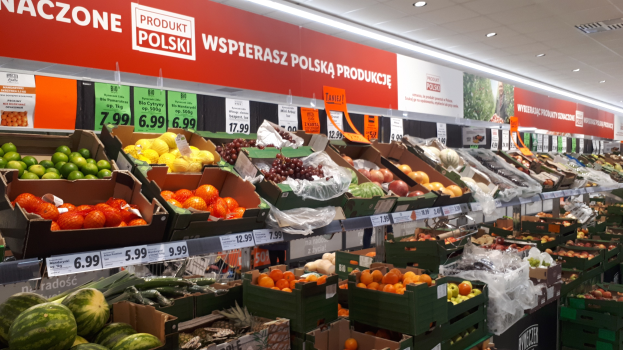 Ein Geschäft mit verschiedenen Obst- und Gemüsesorten in Kartons, jeweils mit Preisschildern, Wandtafeln mit Text und Bildern und Deckenbeleuchtung.