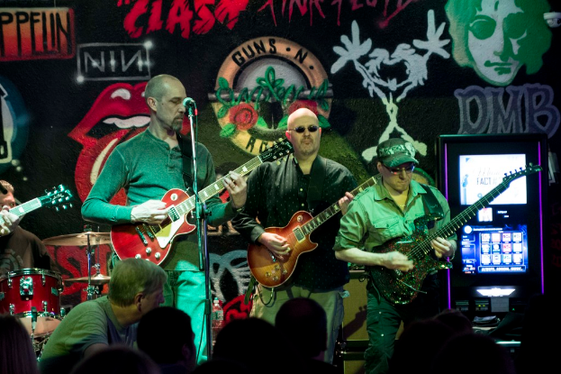 Drei Männer auf einer Bühne bei einem Konzert, mit einer Menge darunter und einer gemalten Wand im Hintergrund.