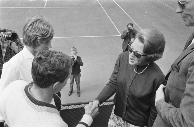Eine Gruppe von Menschen auf einem Tennisplatz, mit einer Frau in einer Brille, die einem Mann im Anzug die Hand schäkelt, und Fotografen im Hintergrund, alles in Schwarz-Weiß.