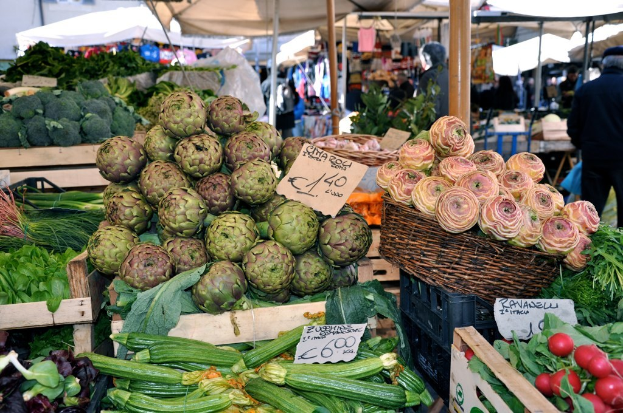 Körbe gefüllt mit Obst, Gemüse und kleinen Tafeln mit Text, mit Zelten und wenigen Menschen im Hintergrund.