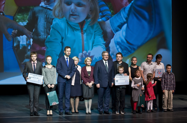 Gruppe von Menschen auf einer Bühne mit Taschen und Urkunden, im Hintergrund ein Bildschirm mit einem Bild eines Kindes und anderen.