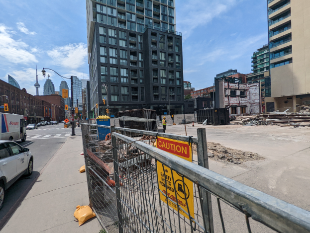 Eine Baustelle mit einem Zaun und einem Schild, das laufende Arbeiten anzeigt, umgeben von Gebäuden, Straßeninfrastruktur, Fahrzeugen, Menschen und Baustoffen, mit einem neuen Wohngebäude im Vordergrund und einem bewölkten Himmel im Hintergrund.