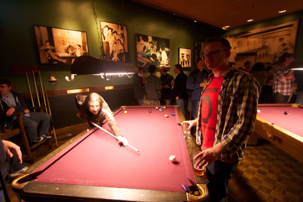 In einem Club spielen zwei Personen Snooker an einem von zwei Tischen, während mehrere andere im Hintergrund stehen und sprechen, vor einer cremefarbenen Wand mit ein paar Fotorahmen.