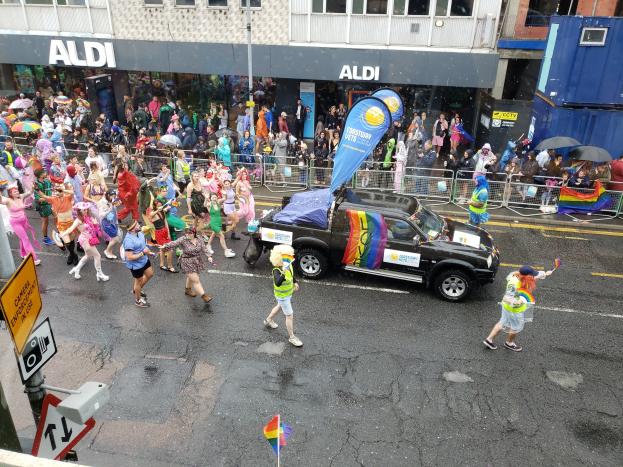 Eine Gruppe bunt gekleideter Menschen, die auf einer Straße in einer Parade gehen, mit einem Auto in der Mitte, einige halten Schirme, und Gebäude im Hintergrund.