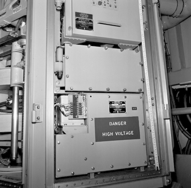 Schwarzes und weißes Foto einer elektrischen Schalttafel mit Warnschild, umgeben von Maschinen und Rohren.