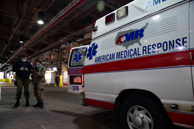 Ein Rettungswagen der American Medical Response (AMR) steht in einem Gebäude mit zwei maskierten Personen davor, ein Fahrzeug auf der linken Seite und mehreren Personen, Lichtern und Rohren im Hintergrund.