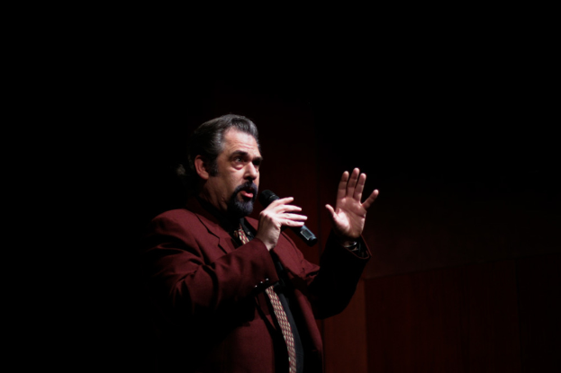 Ein Mann in einem Anzug mit einem Mikrofon in der rechten Hand, der im Dunkeln spricht.