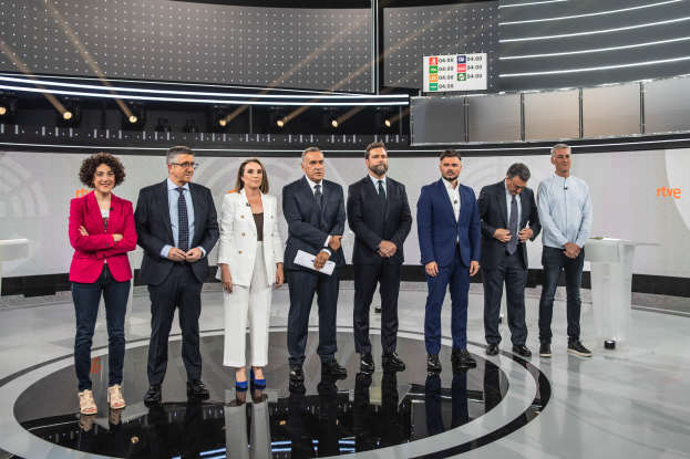 Eine Gruppe von Menschen steht vor einem Fernseher mit zwei Tischen auf jeder Seite, auf denen verschiedene Gegenstände liegen, und einer Wand mit Lichtern und einem Bildschirm im Hintergrund, die an einer Livesendung der spanischen Fernsehshow 'Rive' teilnehmen.