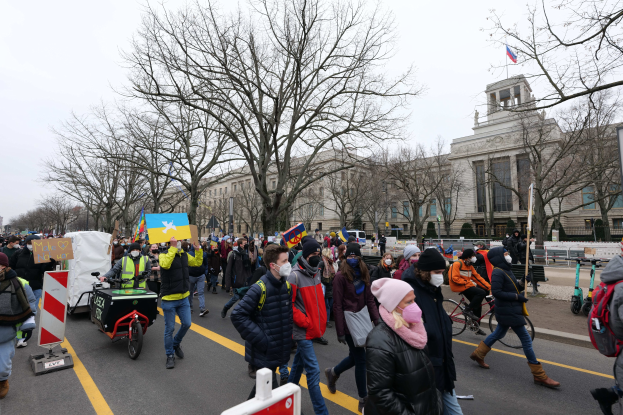 Eine große Gruppe von Menschen marschiert bei einer Demonstration durch eine Straße in Washington, D.C., mit Schildern und Fahrrädern, vor einem Baum, Schildern und einem klaren blauen Himmel im Hintergrund.