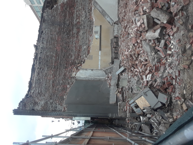 Ein teilweise abgerissener Bau mit verstreuten Ziegeln und Metallstangen, mit anderen Gebäuden und einem klaren blauen Himmel im Hintergrund.