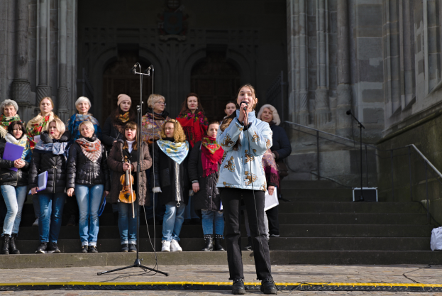 Eine Gruppe von Menschen, einige mit Musikinstrumenten, steht vor einem Gebäude mit Treppen und Geländern, während eine Frau in der Mitte in ein Mikrofon singt, das von Mikrofonstativen umgeben ist.