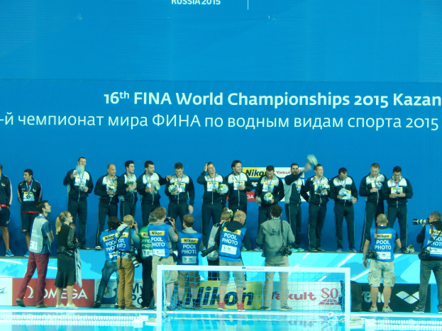 Eine Gruppe von Menschen steht auf einem Swimmingpool, einige halten Kameras, mit einem Netz im Vordergrund und einer Flagge im Hintergrund, auf der steht "FINA World Championships 2015 Kazan".